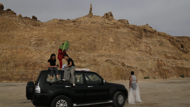 Three girls are sitting on top of a black car. One is holding a large green leaf to cover her face. A man is walking towards them wearing a white skirt. The site of Lot’s wife is in the background.