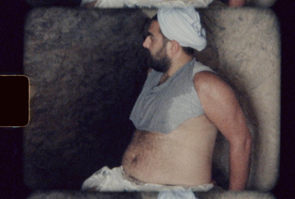 A young man is sitting in Lot’s cave looking ahead. He has a black beard and he is wearing a turban, a cropped top. His stomach is exposed.