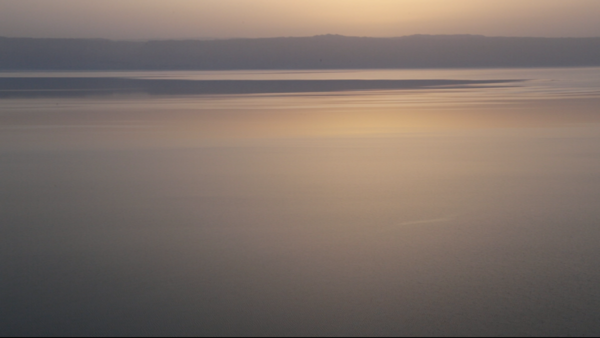 An image of the sunset over the Dead Sea. The mountains in the horizon are purple and the surface of the water and the sky are yellow, pink and blue.