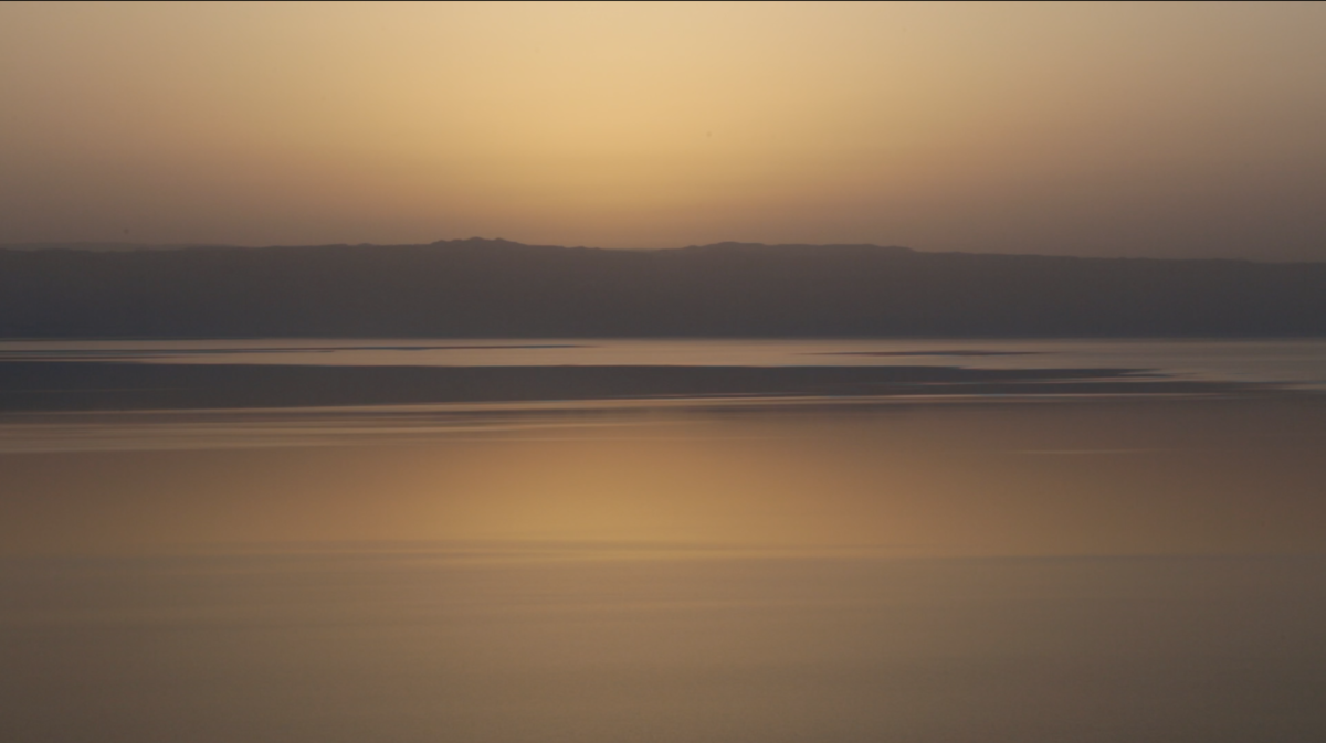 An image of the sunset over the Dead Sea. The mountains in the horizon are purple and the surface of the water and the sky are yellow, pink and blue.