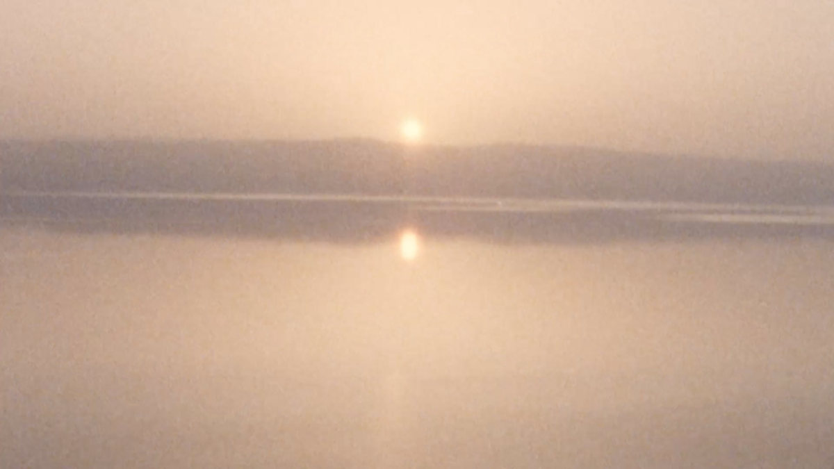 An image of the sun setting over the Dead Sea. The mountains on the horizon and the sun are reflected on the surface of the water.