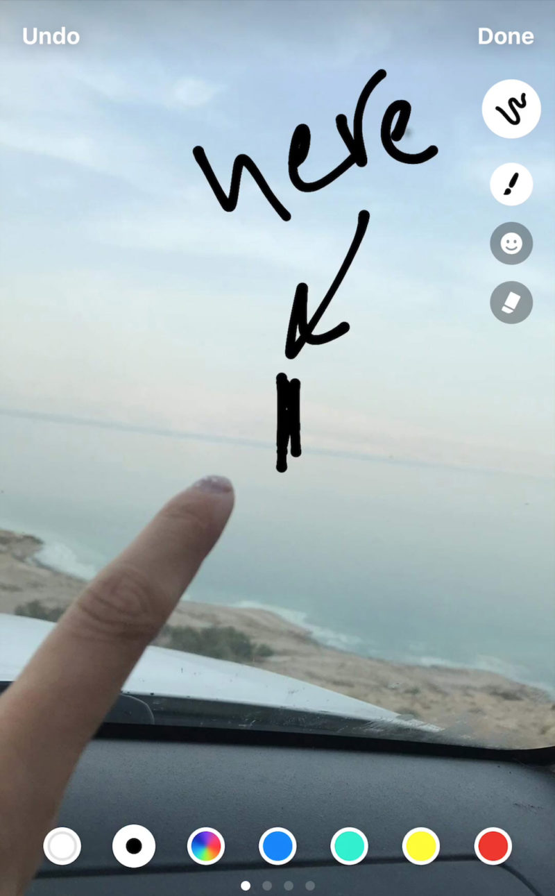 An iphone image of a finger pointing across the Dead Sea to the land visible on the opposite side. There is a hand written word in English and it says “Here".