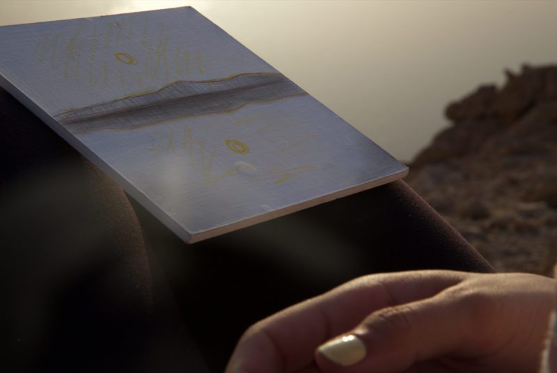 A drawing of a sunset on the Dead Sea. Part of a woman's hand is visible in the corner of the frame.The sea is visible in the background.