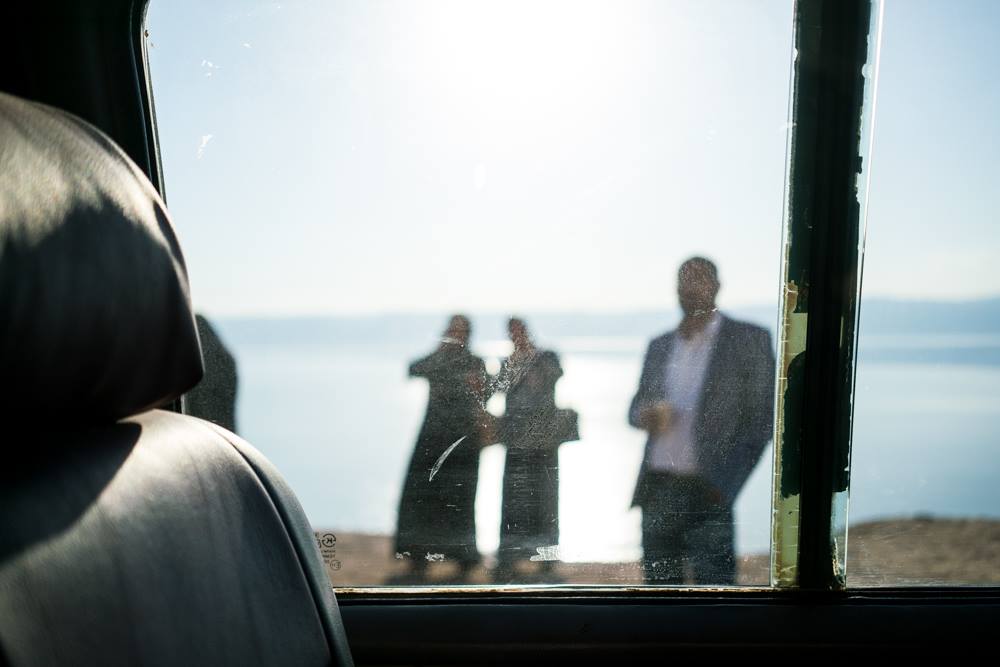 Three bodies stang outside of a bus with the dead sea in the background. The camera is inside the bus and is looking at them through the glass window.