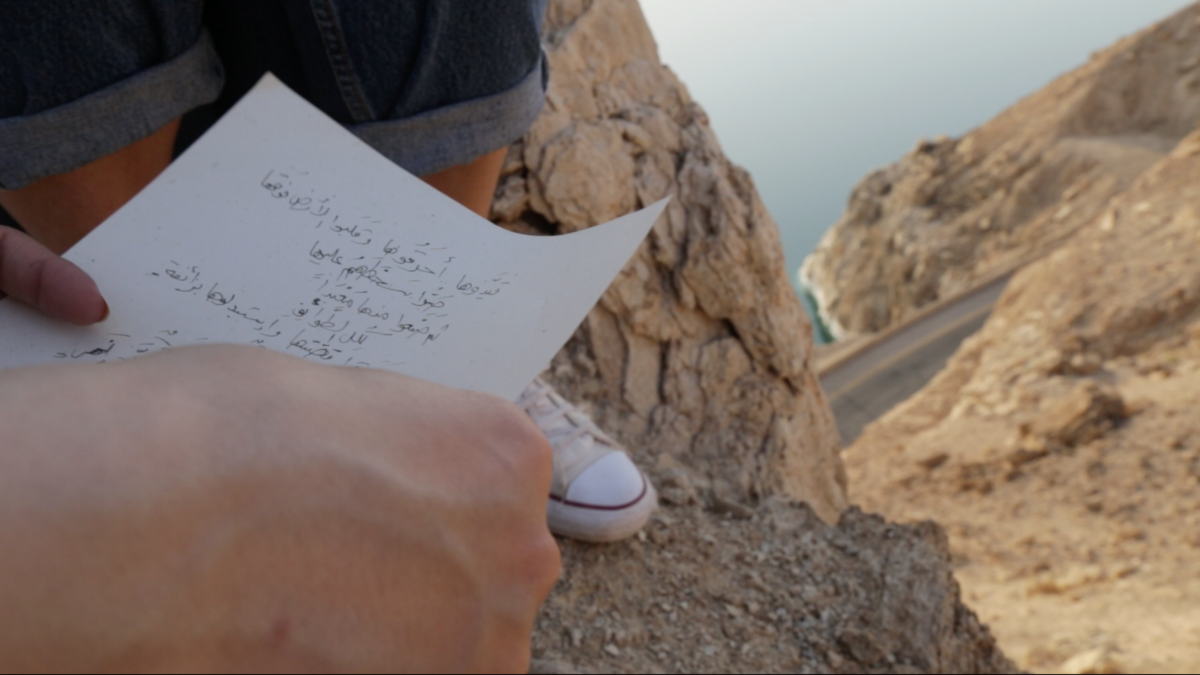 A hand is holding a piece of paper with handwritten arabic text . The person holding the paper is sitting on a rock with