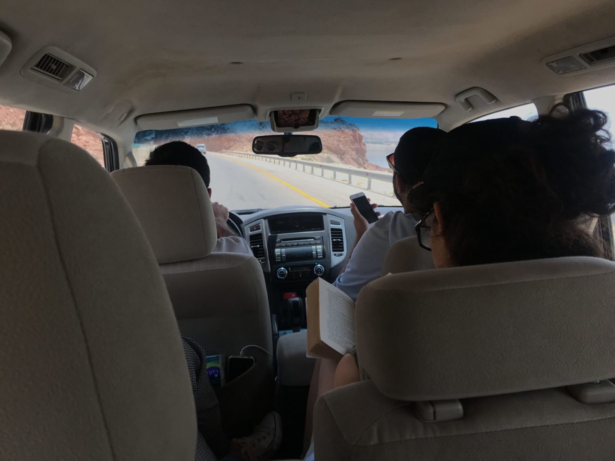 Two men and a woman are inside a car driving on a highway. The woman is sitting in the back seat and she is reading a book.